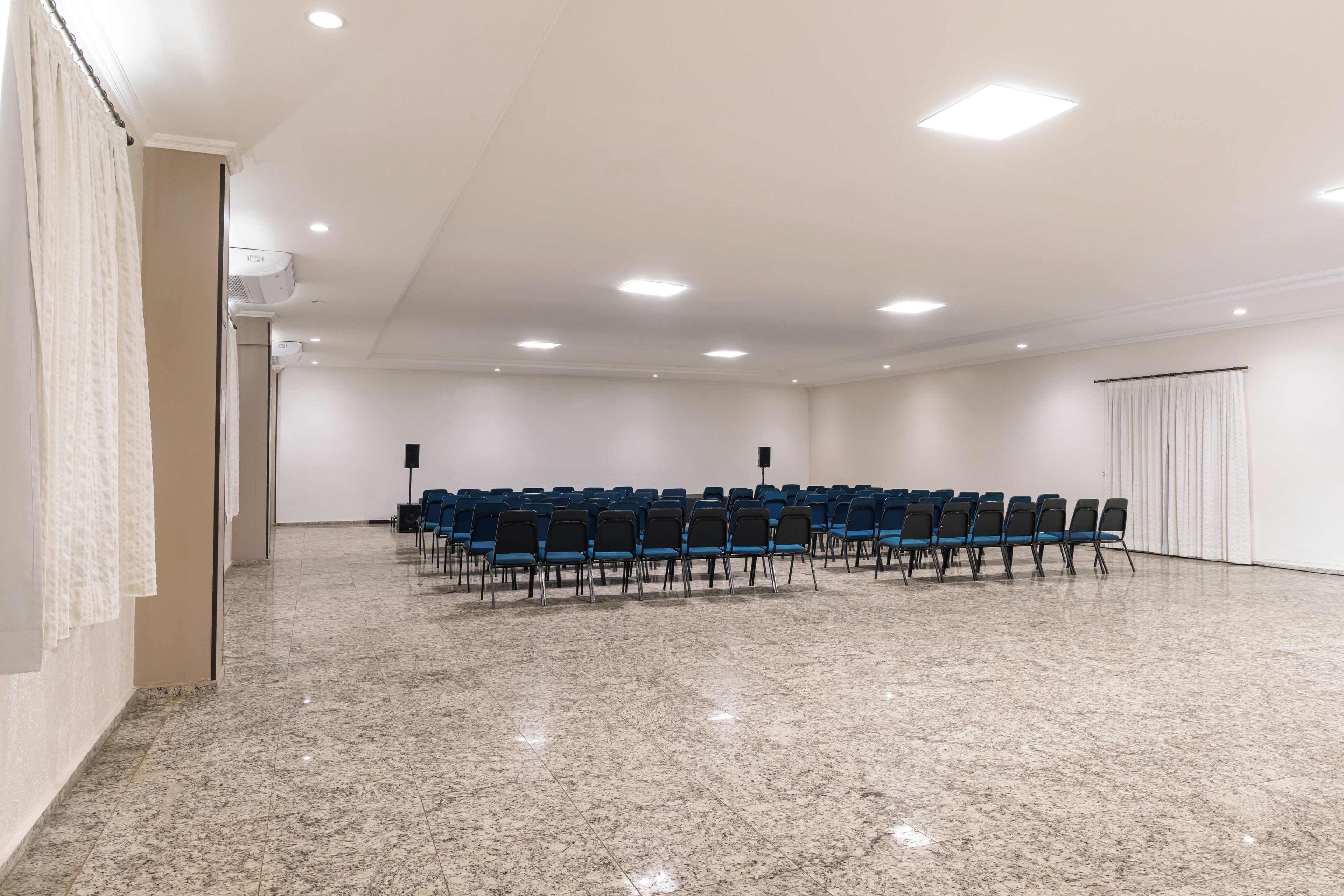 Un salón de conferencias con filas de sillas, techo blanco con iluminación empotrada y una puerta a la derecha.