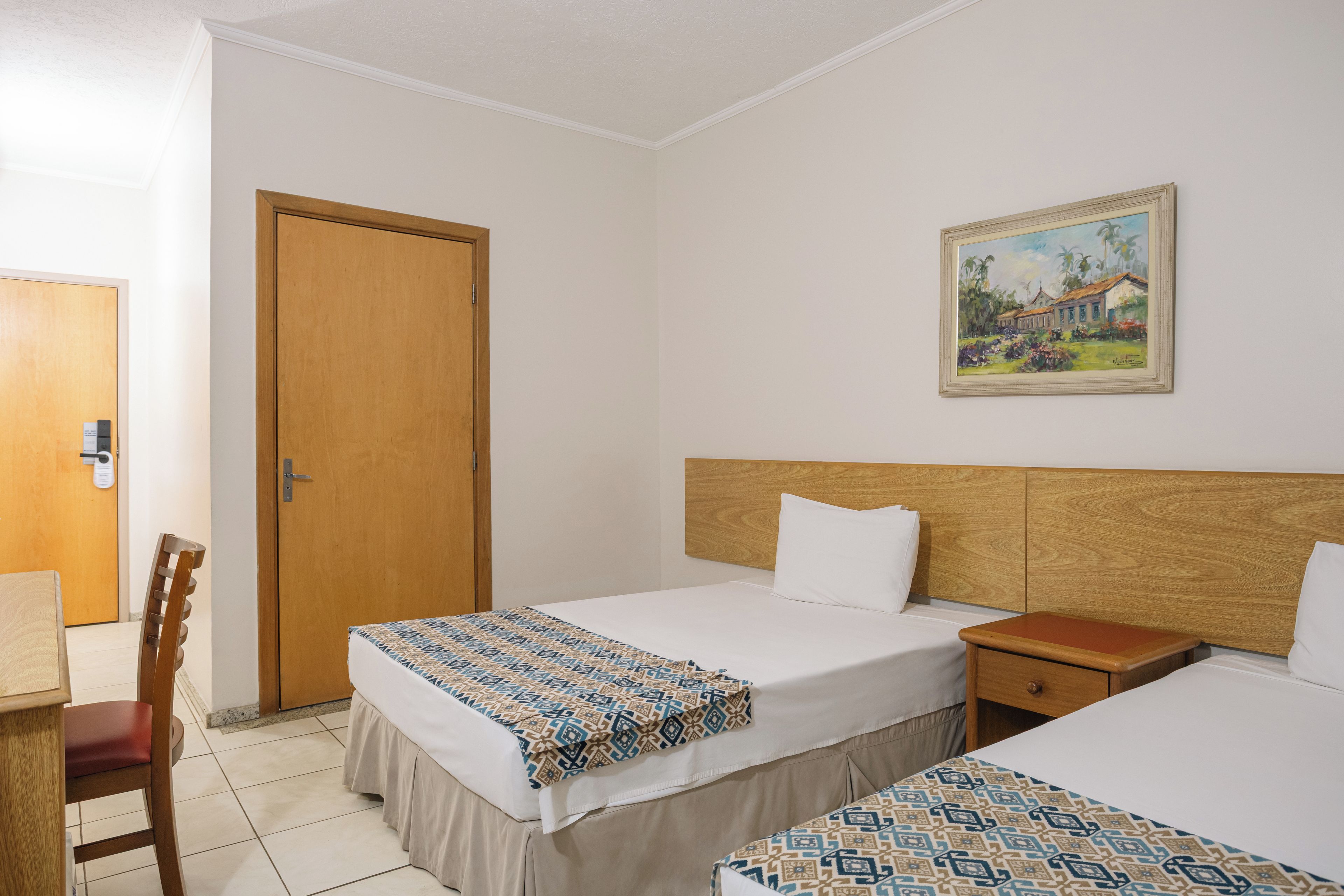 A hotel room with two beds, a wooden door, a painting on the wall, and a chair.