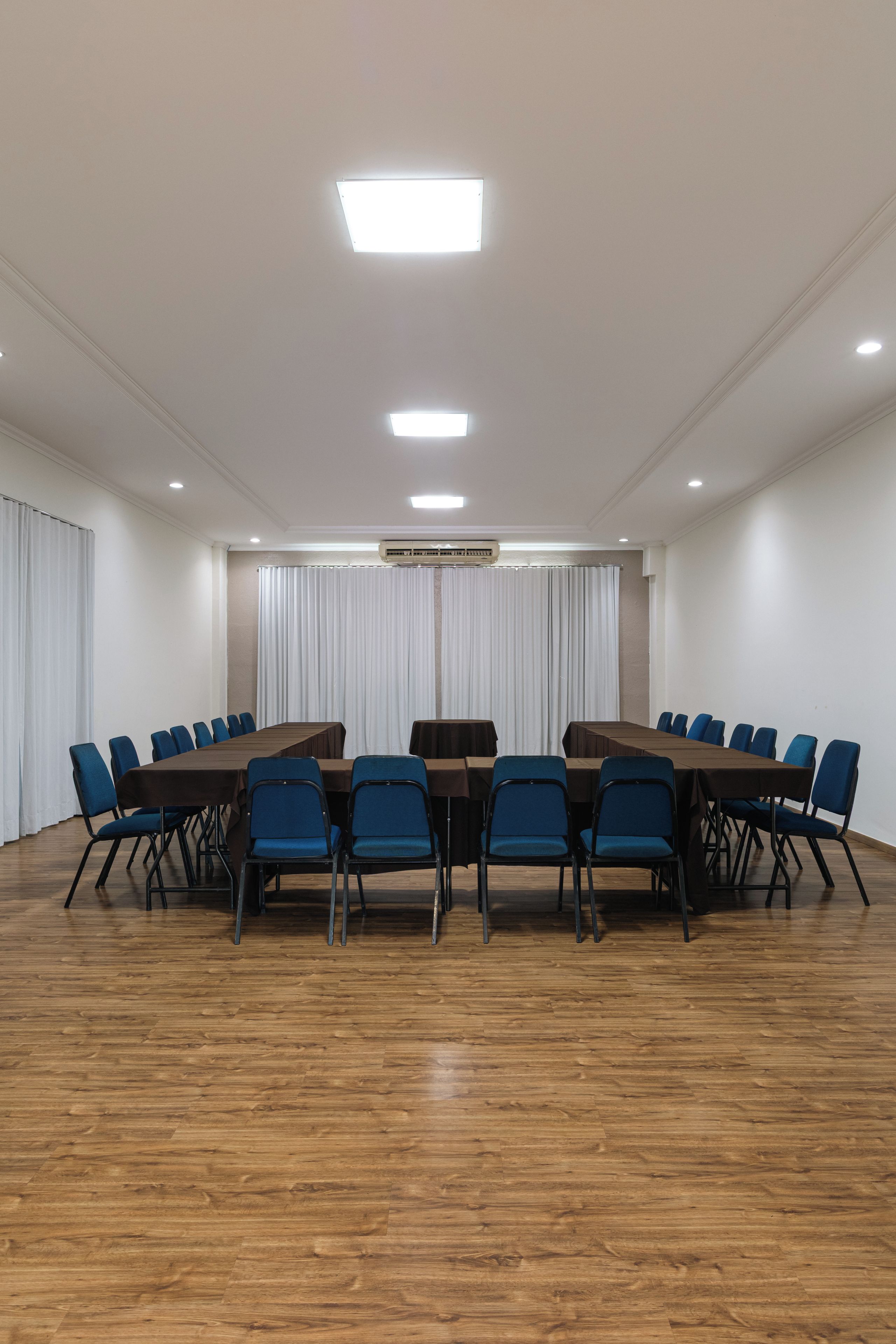 A conference room with a long table, blue chairs, white curtains, and an air conditioning unit on the ceiling.