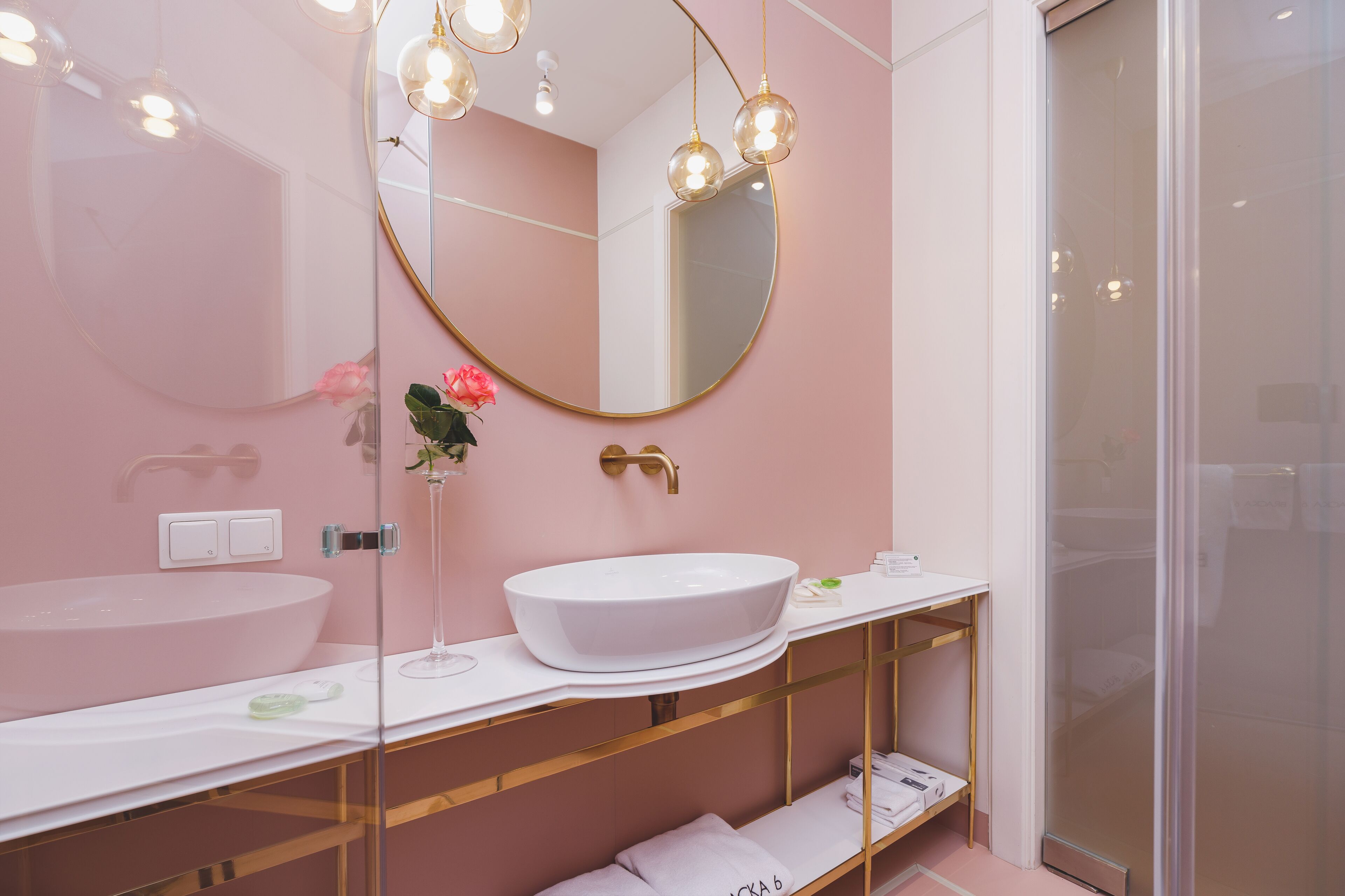 Une salle de bain moderne avec un lavabo blanc, un grand miroir et un autre miroir, plus petit, encadré de doré, placé au-dessus. Deux luminaires sont fixés au-dessus du miroir et, à droite, on aperçoit une cabine de douche en verre.