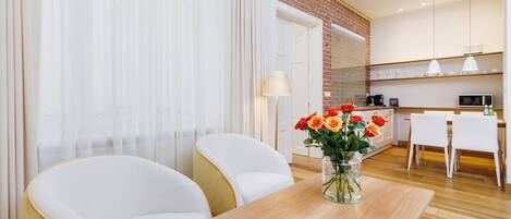 A hotel room with a wooden coffee table, two white armchairs, a vase with flowers, and a kitchen area in the background.