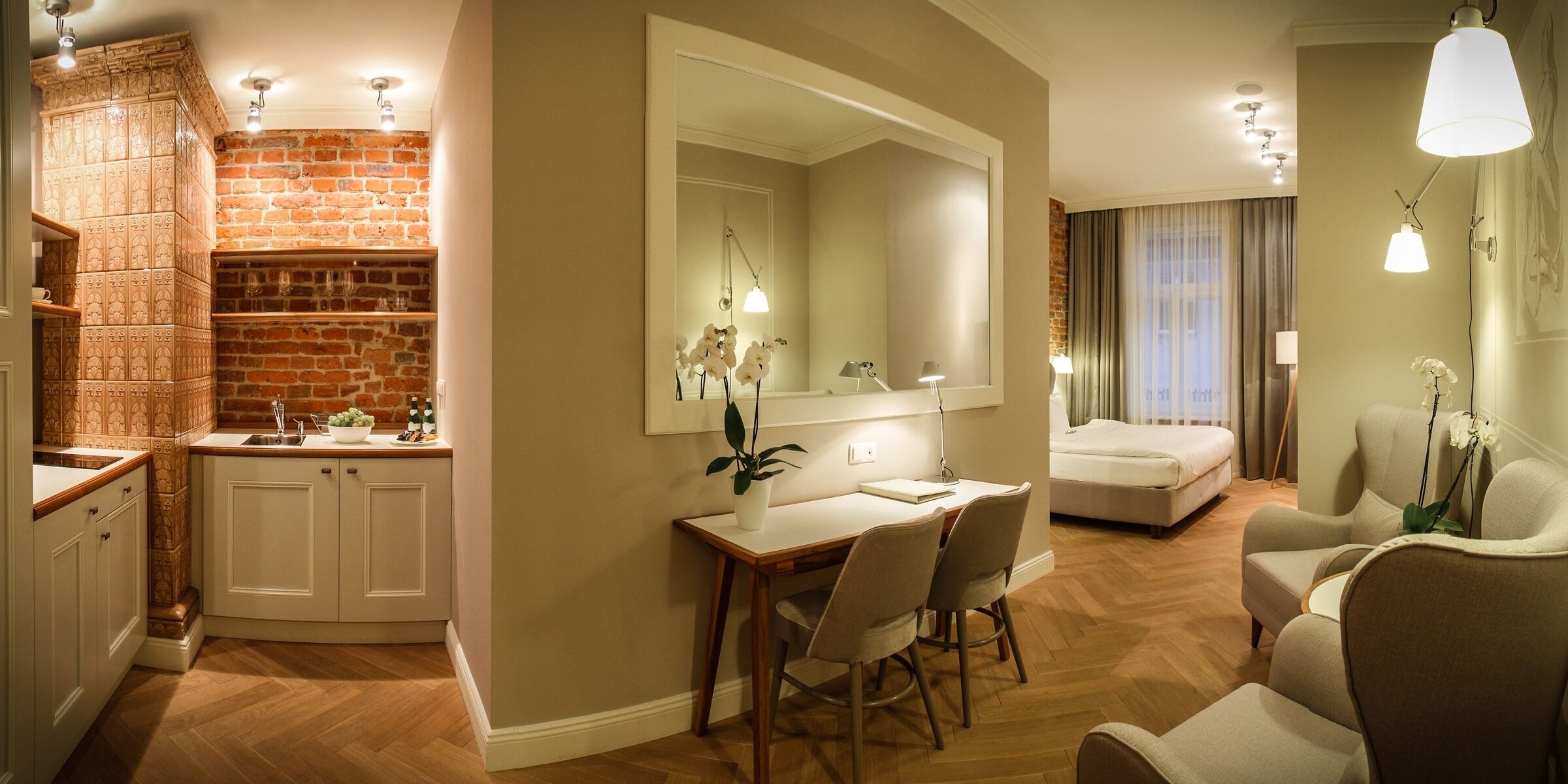 A hotel room with a brick wall, a dining area with a white table and chairs, a large mirror, and a bed with a view of the outdoors.