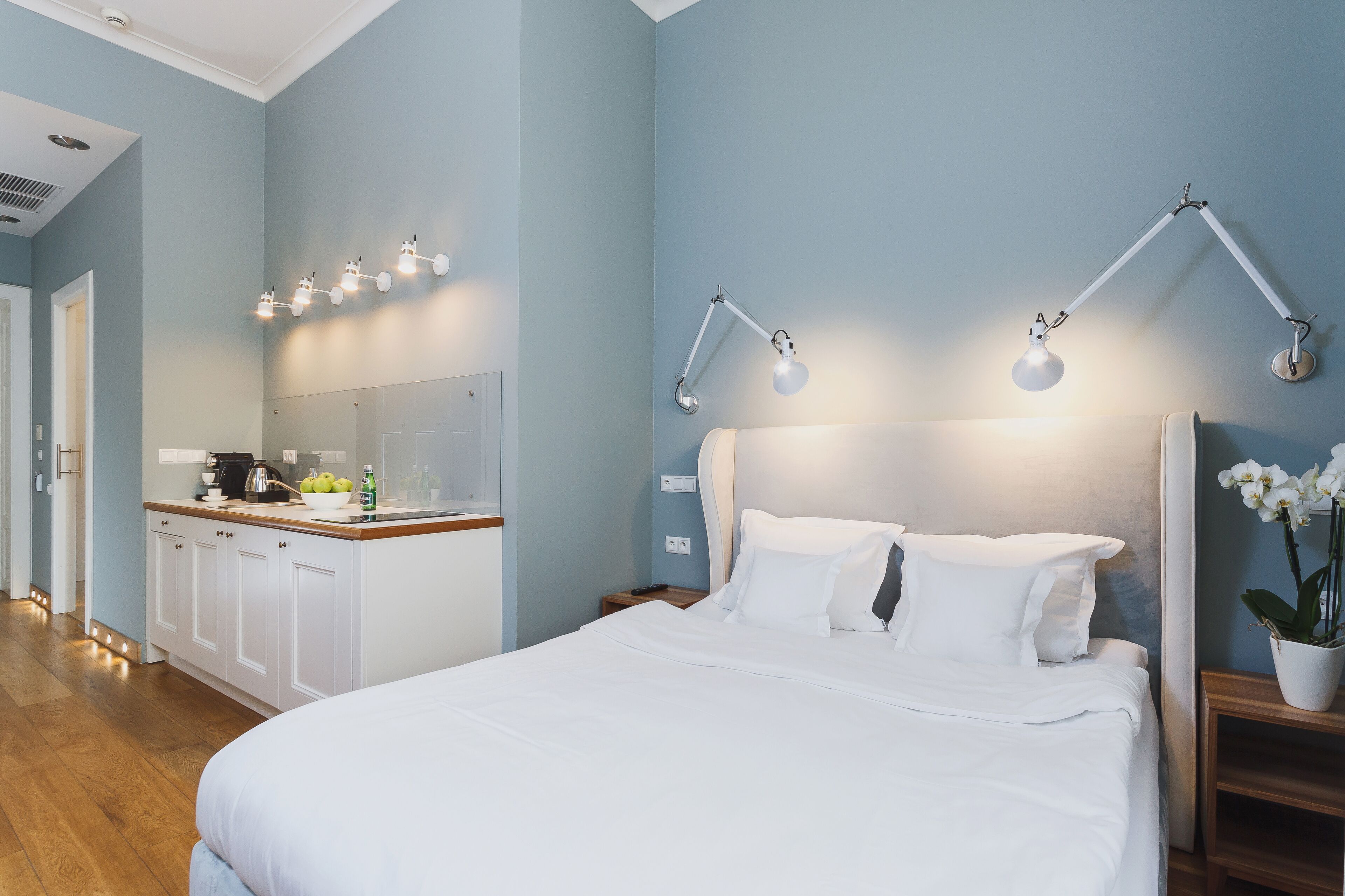 A hotel room with a bed, bedside tables, a nightstand, a desk with a kettle, and a mirror.
