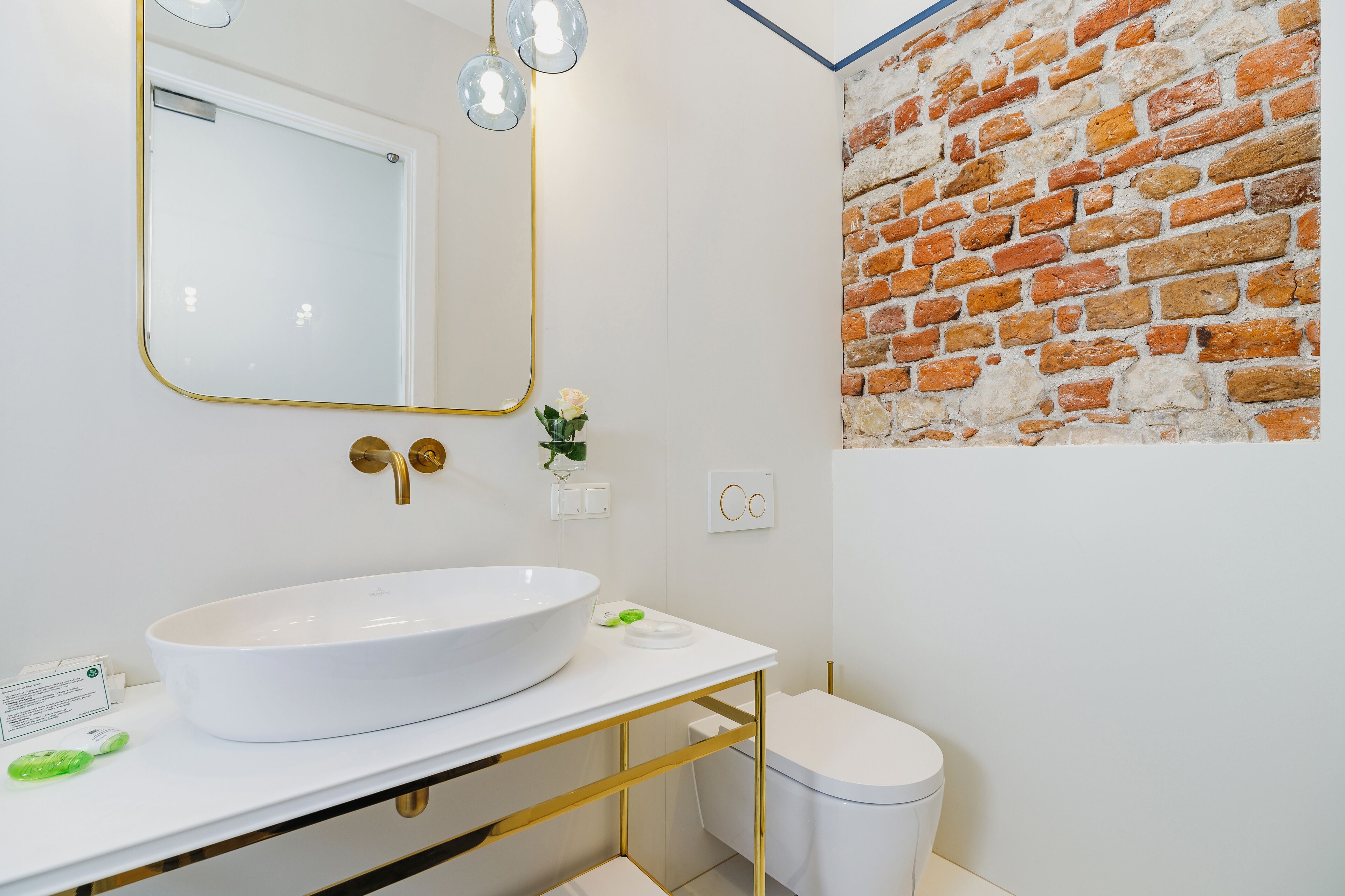 Un baño con un lavamanos blanco, detalles dorados, un espejo y una pared de ladrillo.