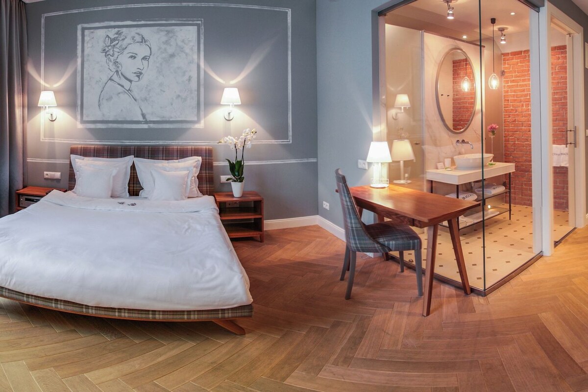A hotel room with a large bed, a desk, a chair, a bathroom with a mirror, and a wall-mounted artwork.