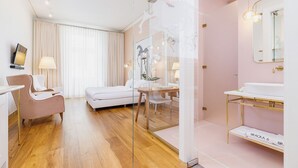 A modern hotel room with a wooden floor, a white sofa, a glass partition, a white desk, a white chair, a white bathroom with a gold vanity, and a large mirror. - Apartamenty Bracka 6 (Kraków)
