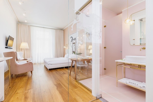 Ein modernes Hotelzimmer mit Holzboden, einer weißen Couch, einer Glastrennwand, einem weißen Schreibtisch, einem weißen Stuhl, einem weißen Badezimmer mit goldfarbener Waschtischgarnitur und einem großen Spiegel.