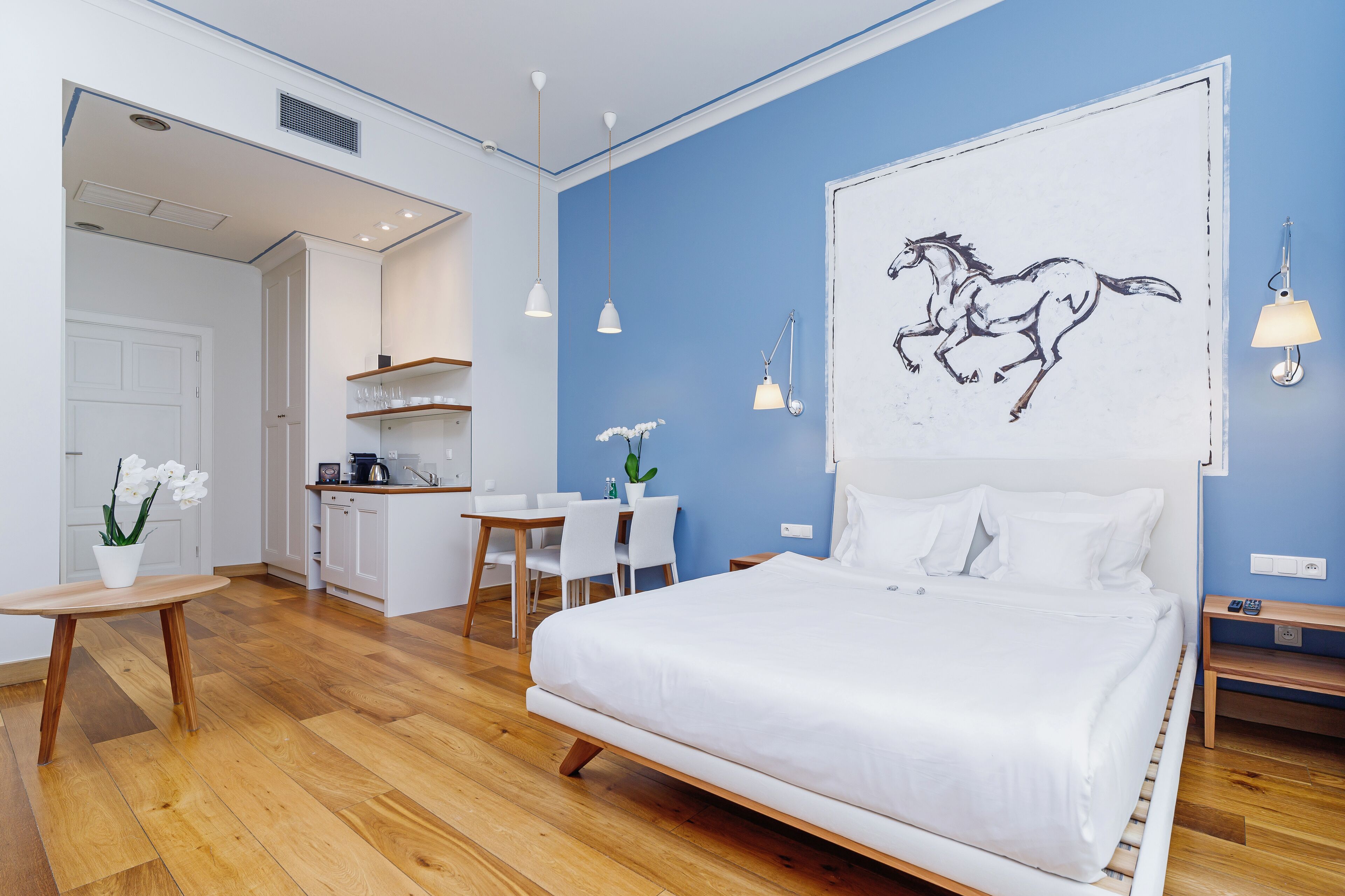 Una habitación de hotel moderna con una cama grande, un escritorio, una silla, una pequeña mesa con flores y una zona de cocina al fondo.