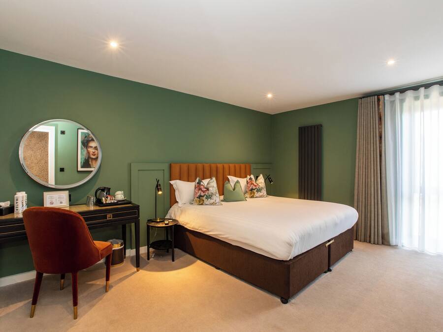 A hotel room with a large bed, a desk with a chair, a round mirror, and a nightstand.