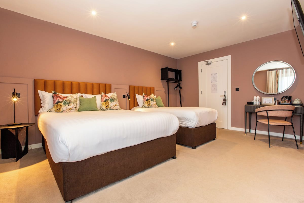 A hotel room with two beds, a desk with a chair, a round mirror, and a small table with a lamp.