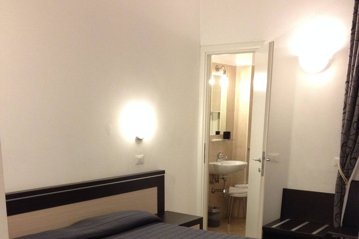 Habitación de hotel con una cama grande, una mesita de noche, un baño con lavamanos y espejo, y zona de ducha.