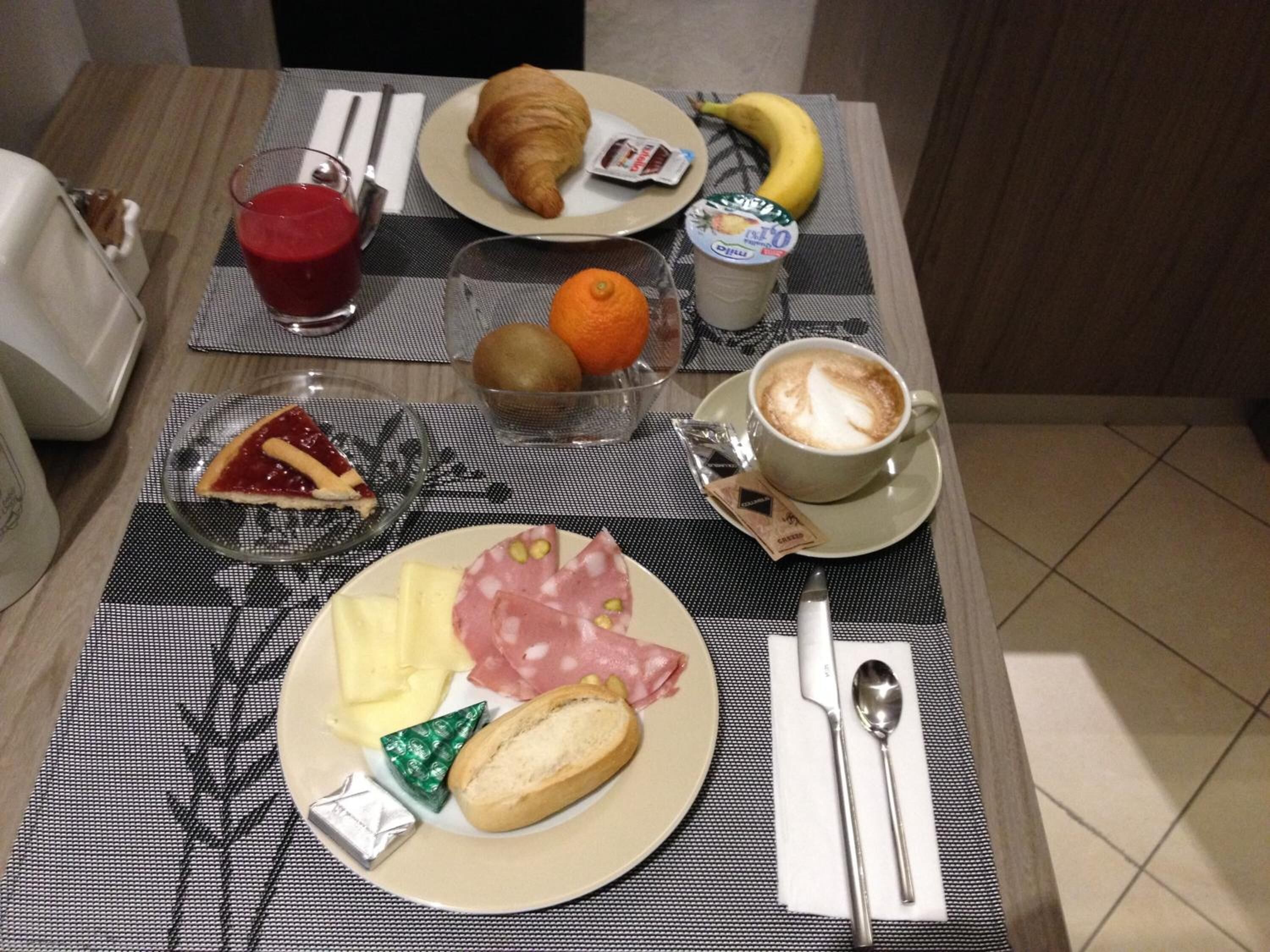 Una mesa de desayuno con croissant, plátano, naranja, kiwi, café, jamón, queso y mermelada.