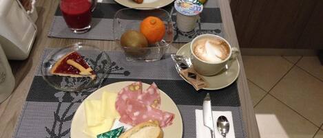Una mesa de desayuno con croissant, plátano, naranja, kiwi, café, jamón, queso y mermelada.