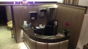 A reception desk with a computer, a phone, and a plant. There are chairs and a small shelf with items on it. - Brenta Milano (Milan)