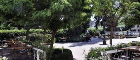 Un área de estar al aire libre con mesas y sillas, rodeada de plantas en macetas y árboles.