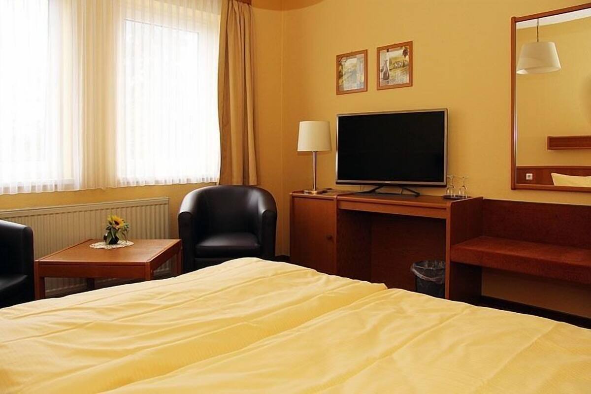 A hotel room with a bed, a desk with a television, a chair, a small table with a vase, and a window with curtains.