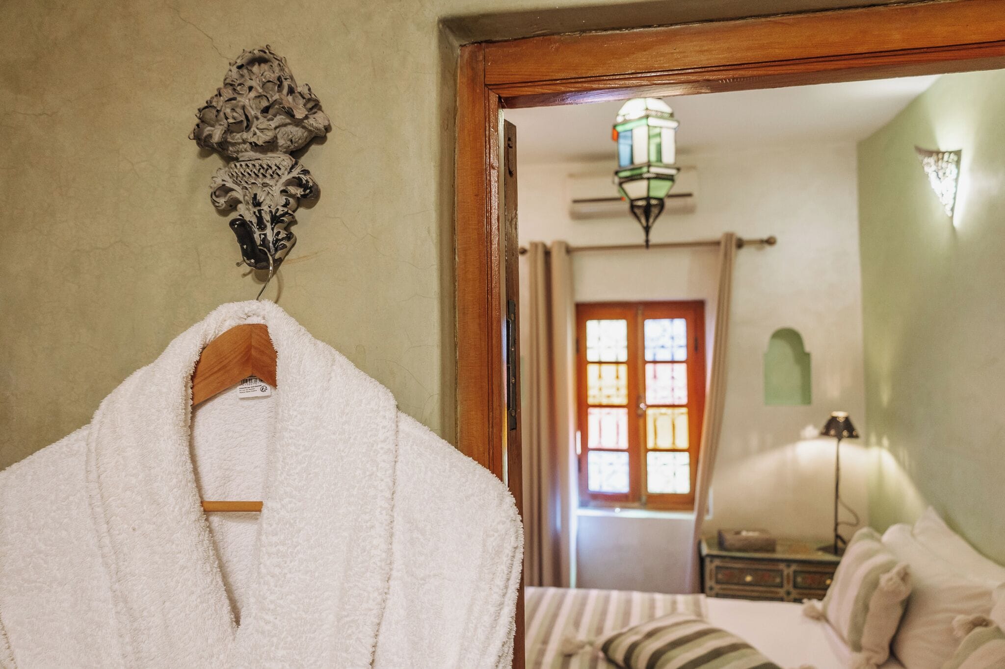 Habitación de hotel con puerta de madera, bata colgada, cama con ropa de cama a rayas, mesita de noche y lámpara fijada a la pared.