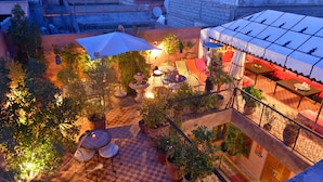 Un espace extérieur aménagé en terrasse, avec un sol à carreaux, plusieurs tables et chaises, des plantes en pot et un grand parasol offrant de l’ombre.