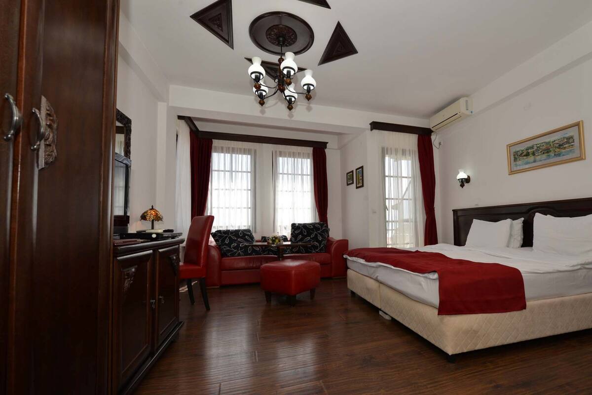 A hotel room with a bed, a desk, a chair, a red ottoman, and a chandelier.