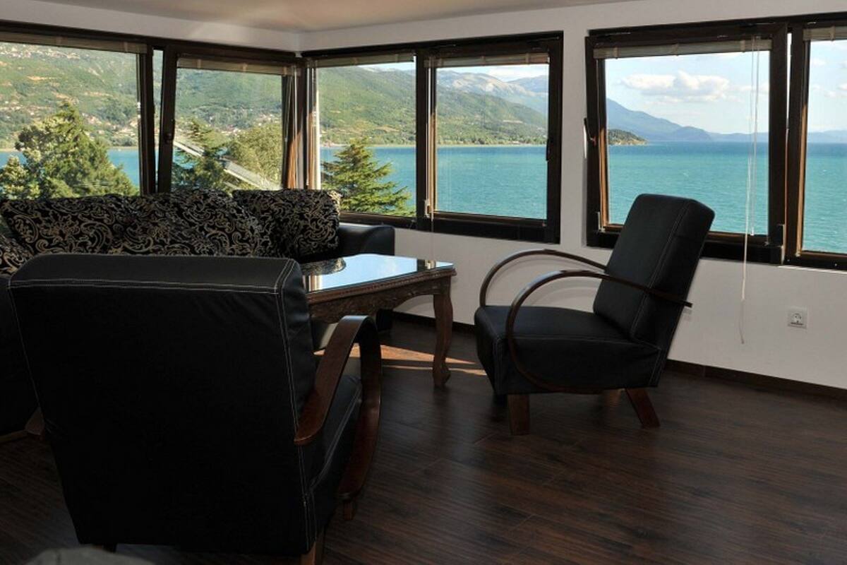 A room with a view of a lake and mountains, featuring a sofa, a table, and a chair.