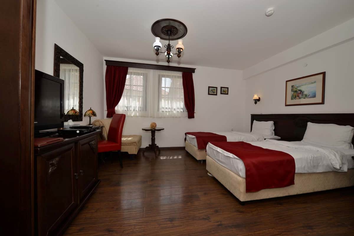 A hotel room with a wooden floor, a TV on a cabinet, two beds with red blankets, a small table with a lamp, and a large mirror.