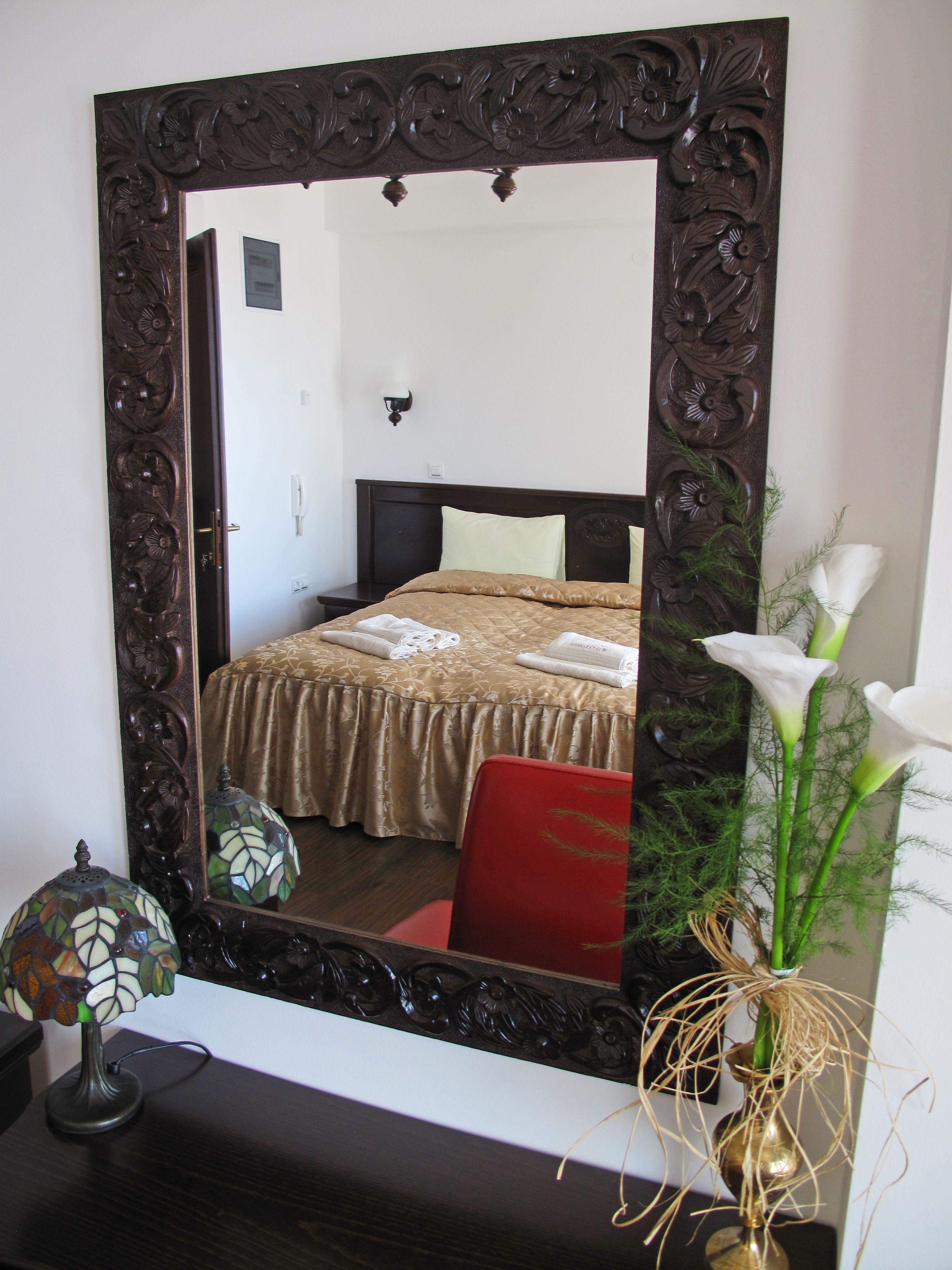 Ein Schlafzimmer mit einem großen, verzierten Spiegel, einem Bett mit goldener Tagesdecke, einem roten Sessel und einer Vase mit weißen Blumen.
