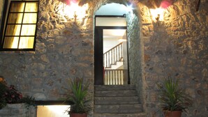 Entrada de un edificio de piedra con una escalera que conduce al interior, plantas en macetas y una ventana con cortinas.