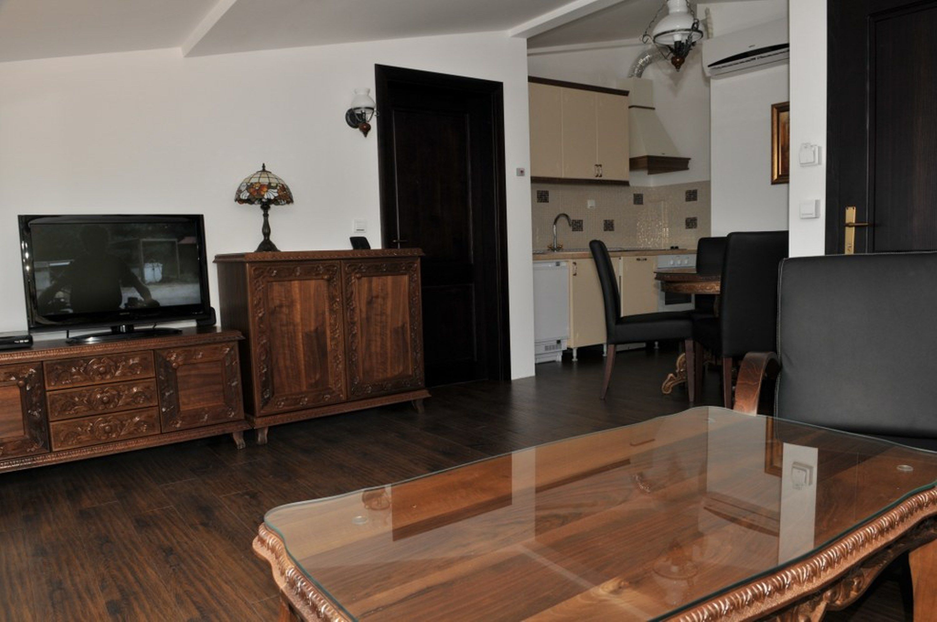 A living room with a wooden cabinet, a glass coffee table, a television, and a kitchen in the background.