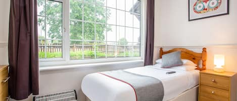 A bedroom with a bed, bedside table, lamp, radiator, and a window with curtains.