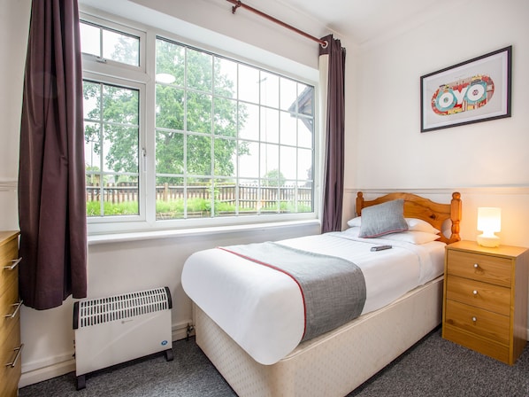 A bedroom with a bed, bedside table, lamp, radiator, and a window with curtains.