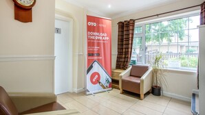 Un espace d’accueil d’hôtel avec une horloge, une banderole rouge faisant la promotion de l’application OYO, un coin salon avec une chaise et un pot de fleurs, et une fenêtre donnant sur l’extérieur.