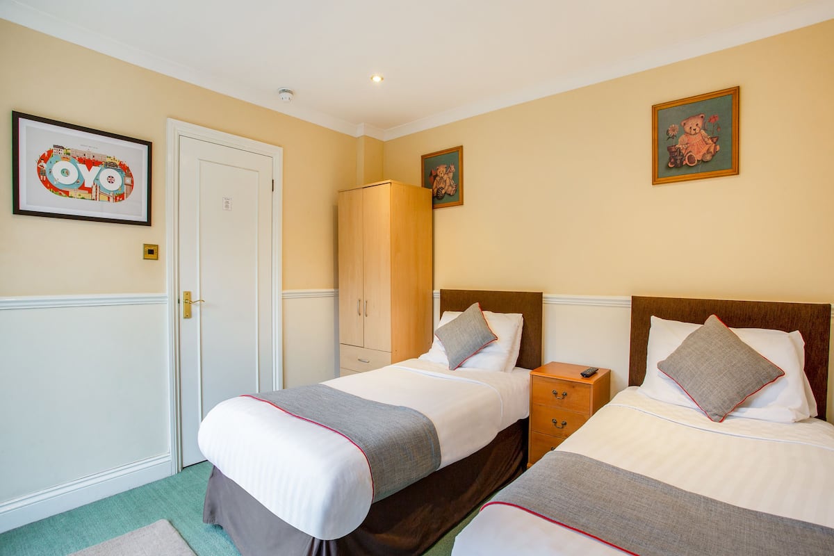 Quarto de hotel com duas camas, um guarda-roupa, uma mesa de cabeceira e um pequeno armário.