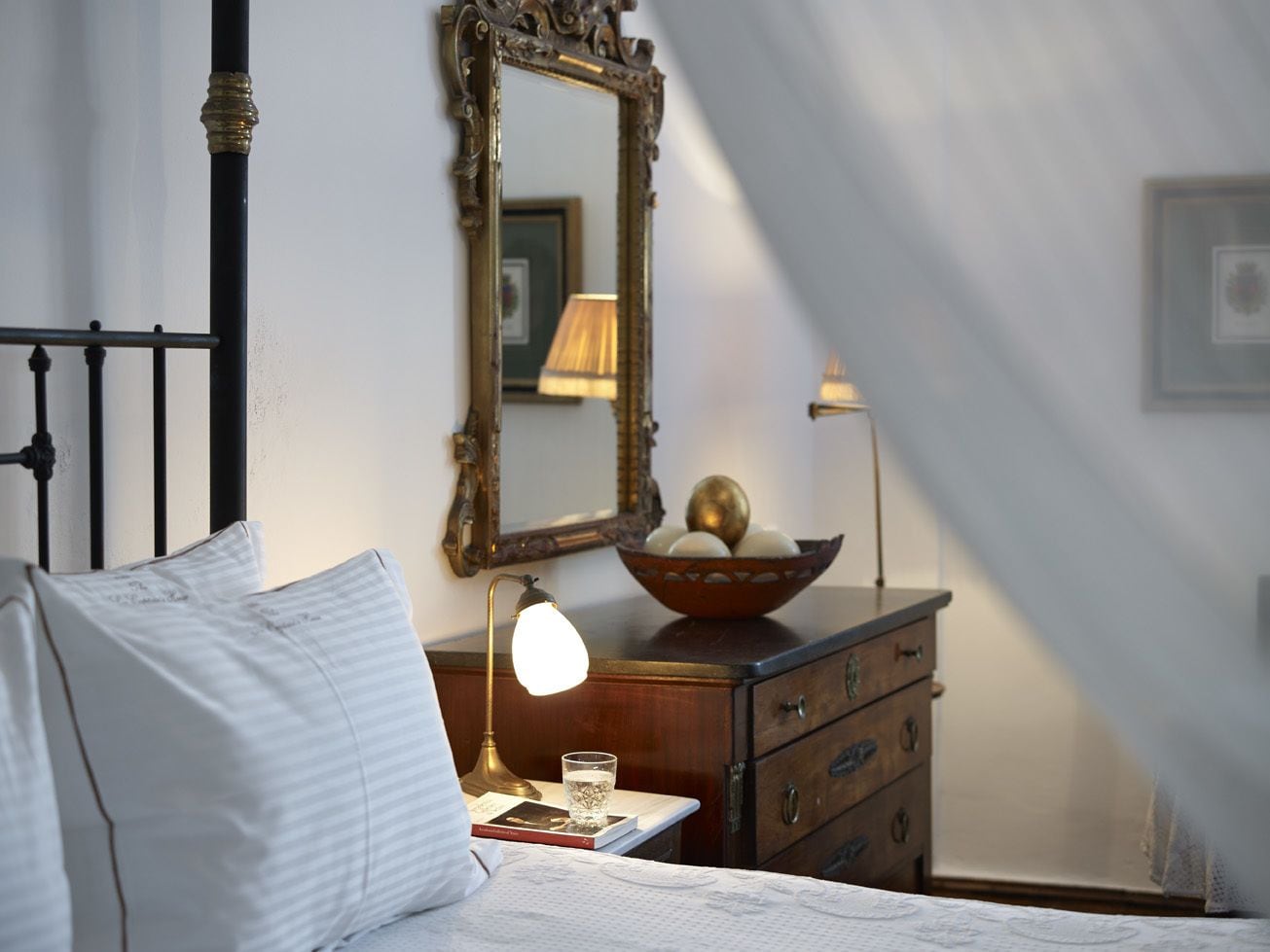 Un dormitorio con cama, mesita de noche con lámpara, cómoda, espejo y un cuadro en la pared.
