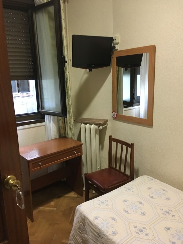 A room with a wooden desk, a chair, a bed with a patterned bedspread, a television mounted on the wall, and a radiator. - Hostal Galaico (Madrid)