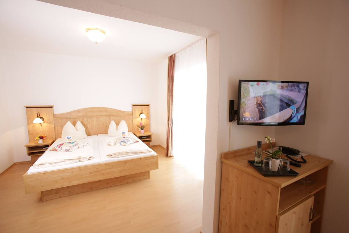 Une chambre d’hôtel avec un lit en bois, deux lampes de chevet, un téléviseur fixé au mur et un bureau en bois agrémenté d’une plante et d’une bouteille.