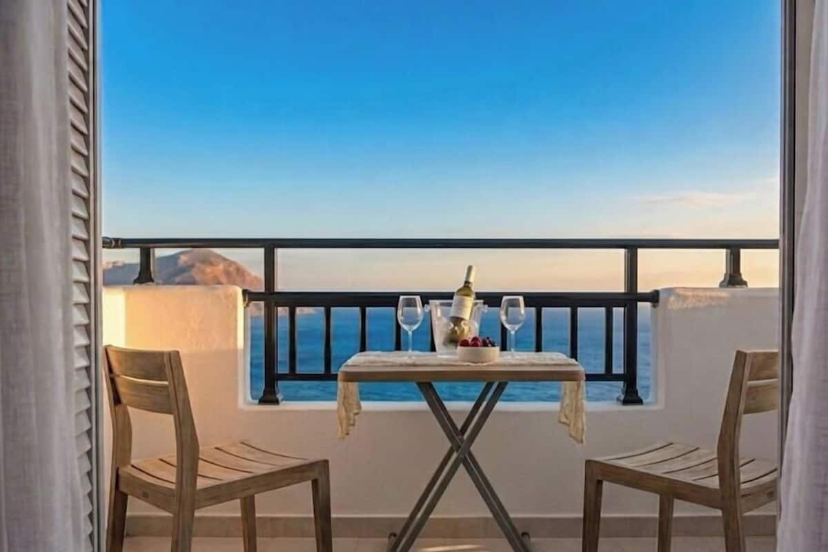 A balcony with a table set for two, a bottle, and glasses, overlooking a clear view of the sea.