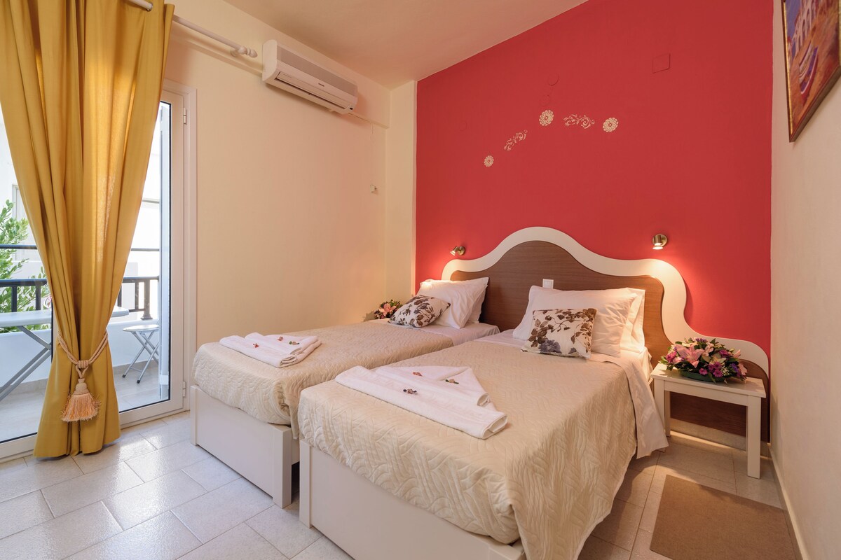 Habitación de hotel con dos camas, una pared de acento roja, un balcón con cortinas amarillas y una pequeña mesa con un arreglo floral.