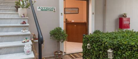 Un área de recepción con una escalera, una planta en maceta, un pequeño árbol y un letrero que dice "ANTIGONI RECEPTION".