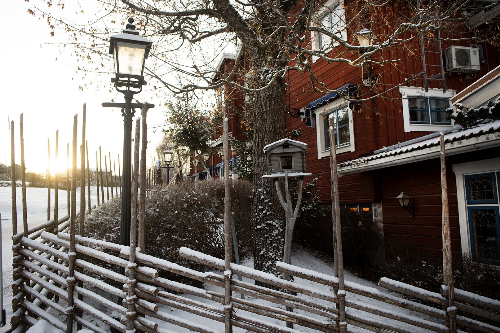 A wooden fence with snow-covered railings, a street lamp, and a birdhouse on a tree.