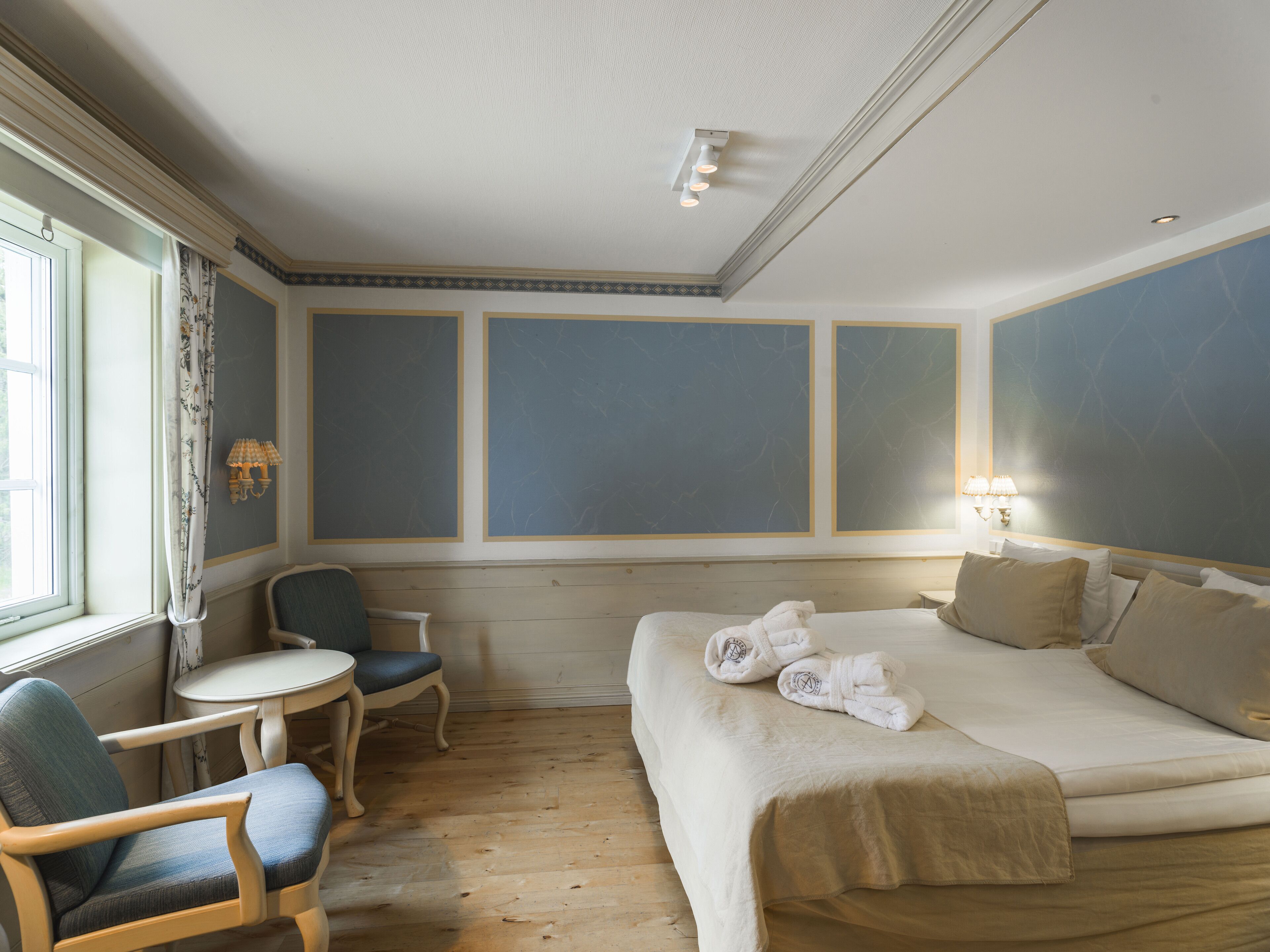 A hotel room with a bed, a chair, a small table, and a window with curtains.