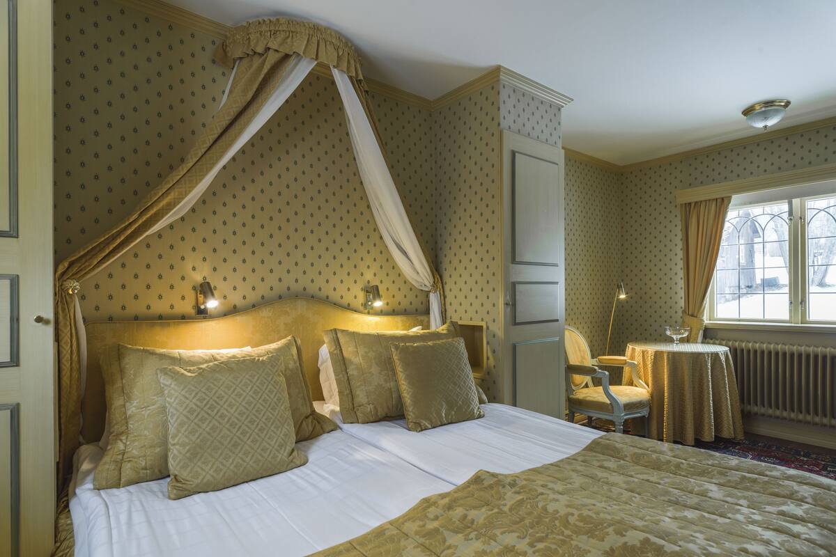 Ein Schlafzimmer mit Himmelbett, Nachttisch, Stuhl und einem fenster mit Vorhängen.