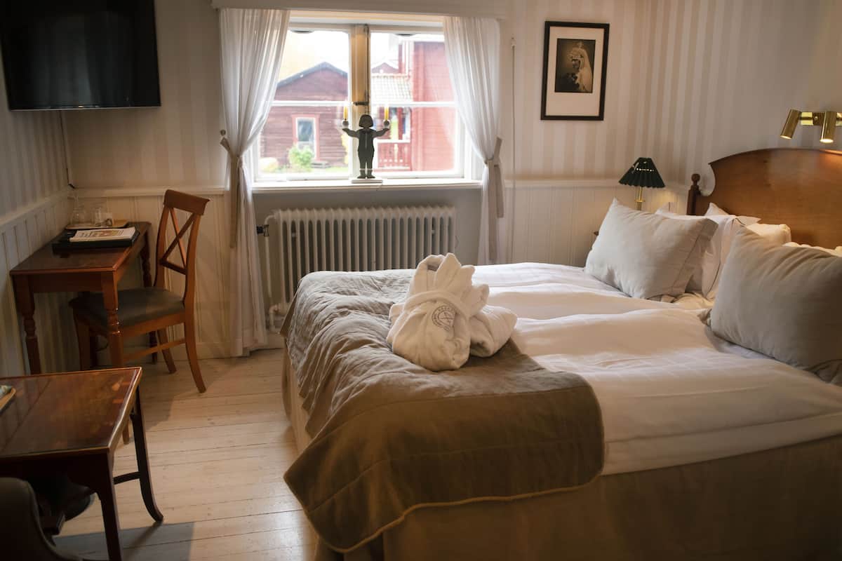 Ein ordentlich eingerichtetes Hotelzimmer mit Bett, Schreibtisch, Stuhl, einem Fenster mit Vorhängen, einem Heizkörper und einem an der Wand befestigten Fernseher.