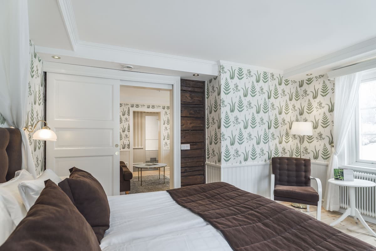 Ein Hotelzimmer mit einem großen Bett, einer Sitzecke mit Stuhl und Tisch, Blickrichtung zum Badezimmer und einer gemusterten Tapete.