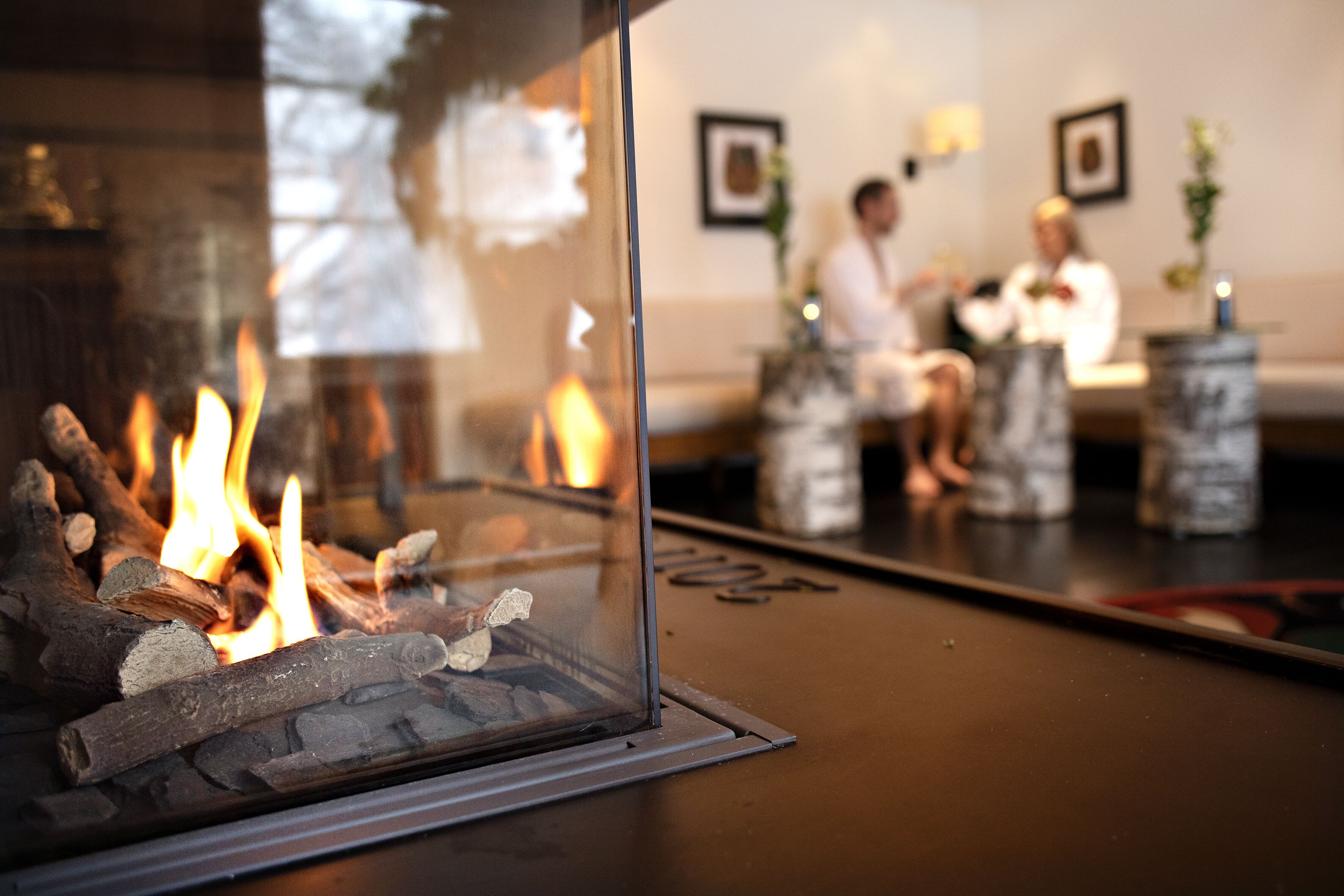 Una chimenea con leña ardiendo, un vidrio y el reflejo de una habitación con un sofá y dos personas.