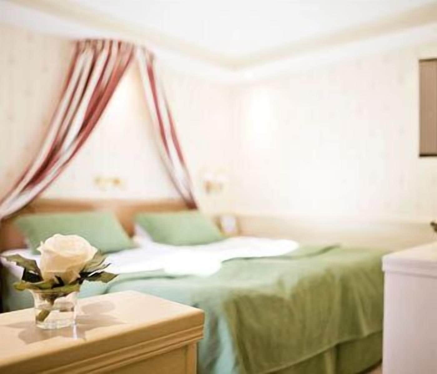 Un dormitorio con cama, ropa de cama verde, una mesita de noche con un florero y una ventana con cortinas.