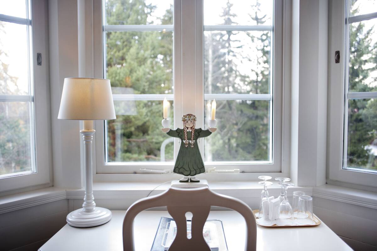 Ein Tisch mit einer weißen Lampe, einer Figur und Kerzenhaltern vor einem Fenster mit Blick auf Bäume.