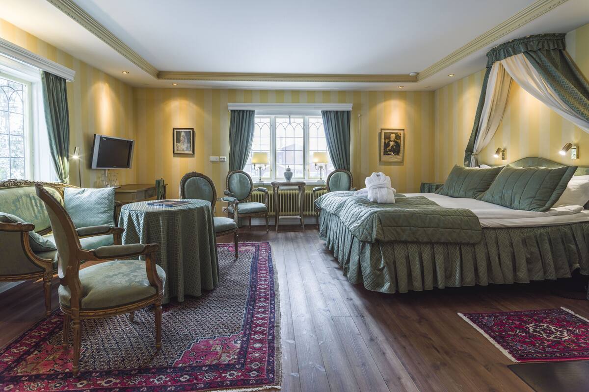 Ein stilvolles Hotelzimmer mit einem großen Bett, einer Sitzecke mit Tisch und Stühlen, einem Fernseher und Blick ins Freie durch ein Fenster.