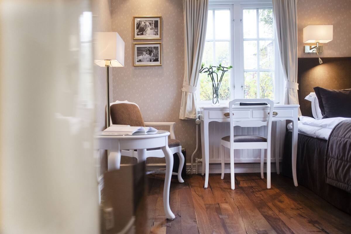 Ein gemütliches Zimmer mit einem Bett, einem Schreibtisch mit Stuhl, einem kleinen Tisch, einer Lampe und einem Fenster mit Vorhängen.