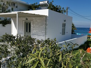 A white building with a door and windows, surrounded by greenery and a view of the sea.