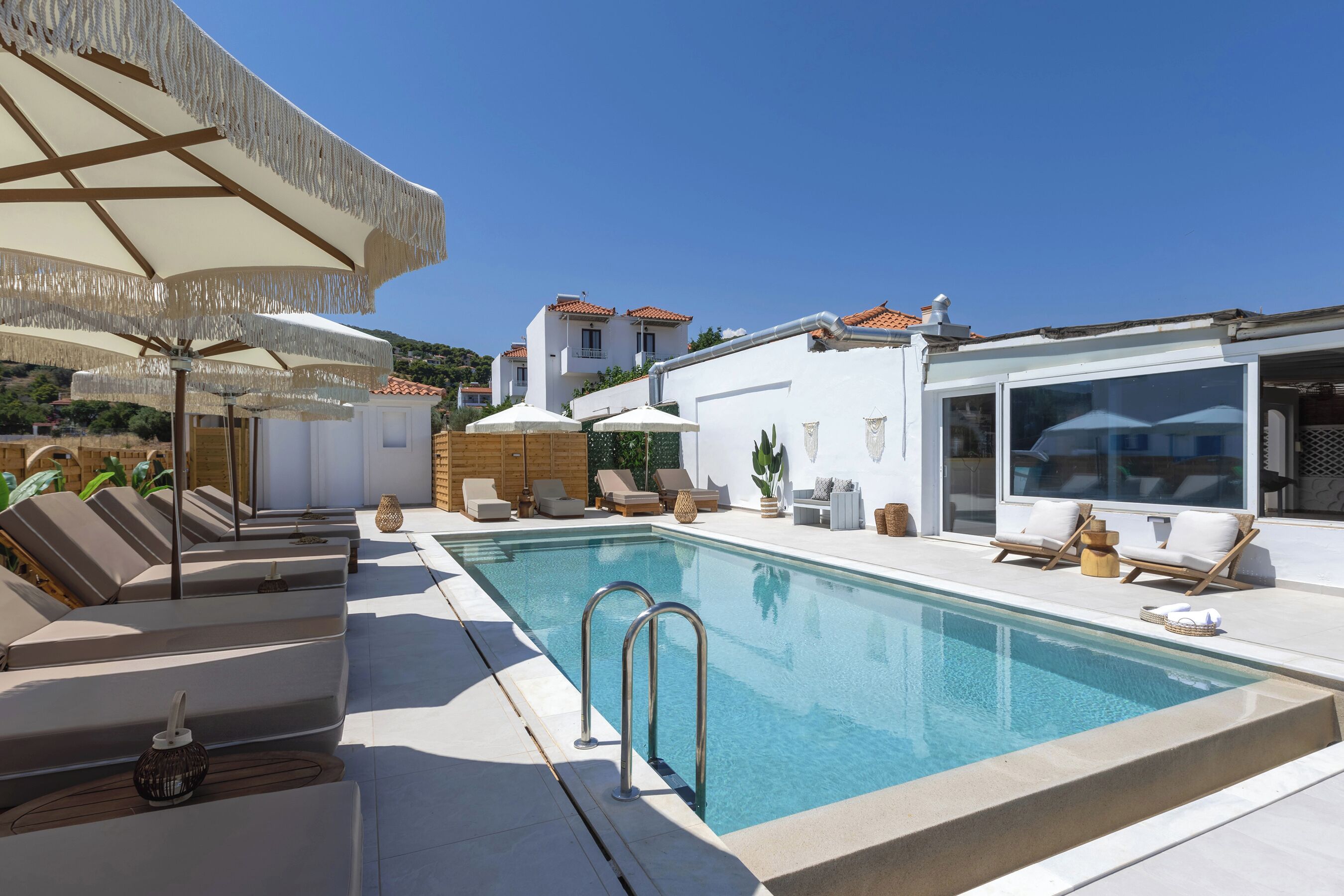 A pool area with lounge chairs and umbrellas, a clear blue pool, and a white building with a tiled roof in the background.